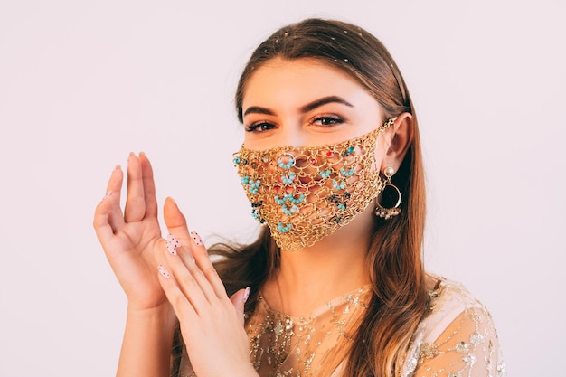 Pandemic party. Holiday greeting. Covid-19 festive look. Happy woman in handmade ethnic style gold chain face mask matching outfit applauding on pastel pink background.