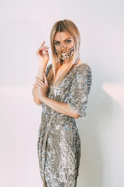 Pandemic party look. Fashion trend 2021. Festive DIY accessory. Mysterious woman in glamour chain face mask matching silver shiny sequin dress on pastel blue pink light background.