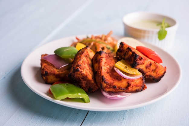 Paneer Tikka or chilli paneer Kabab - Tandoori Indian cheese skewers, served in white plate with colourful capsicum and onion, with green sauce