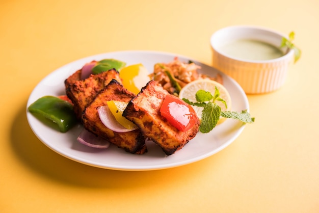 Paneer Tikka of chili paneer Kabab - Tandoori Indiase kaasspiesjes, geserveerd in wit bord met kleurrijke paprika en ui, met groene saus