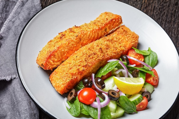Panko crusted baked salmon fillets with spinach tomato cucumber olives salad on a plate on a dark wooden table,  view from above, close-up