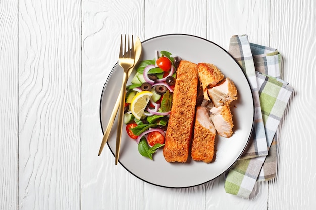 Photo panko crusted baked salmon fillets with spinach tomato cucumber olives salad on a plate on a white wooden table,  view from above, flat lay, free space