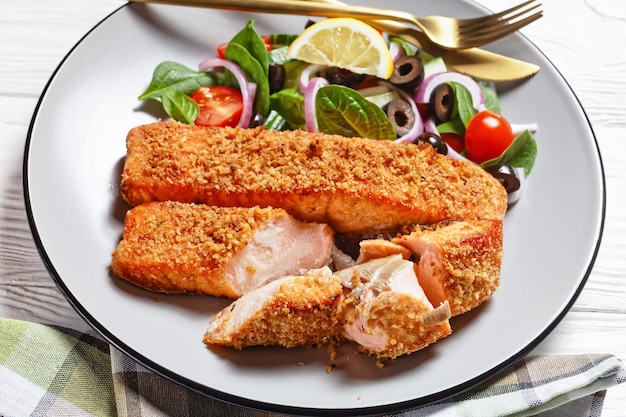 Panko crusted baked salmon fillets with spinach tomato cucumber olives salad on a plate on a white wooden table,  view from above