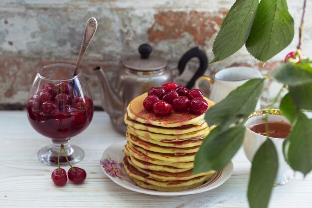 Pannenkoeken en kersenjam staan op tafel met een kopje en een brouwer op de achtergrond van een vintage bakstenen muur