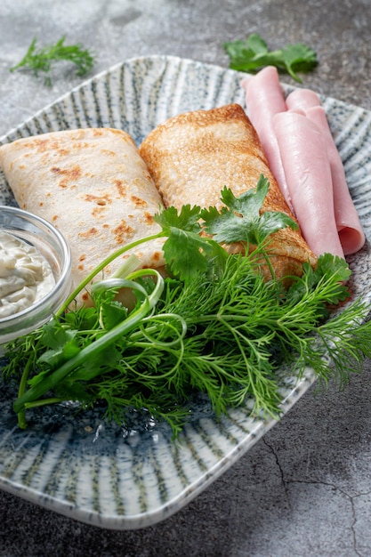 Pannenkoeken gevuld met ham in een bord met peterselie en dillegroen en zure room op een grijze stenen tafel, flatlay