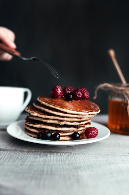 Pannenkoeken met bessen en honing op witte plaat, hand met vork, lepel in pot, houten tafel, kopje thee. Hoge kwaliteit foto