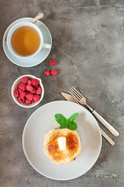 Pannenkoeken met boter. Een blauwe plaat. Afgewerkt met een takje munt. Vlakbij staat een kopje met frambozen en een mok thee. Uitzicht van boven.