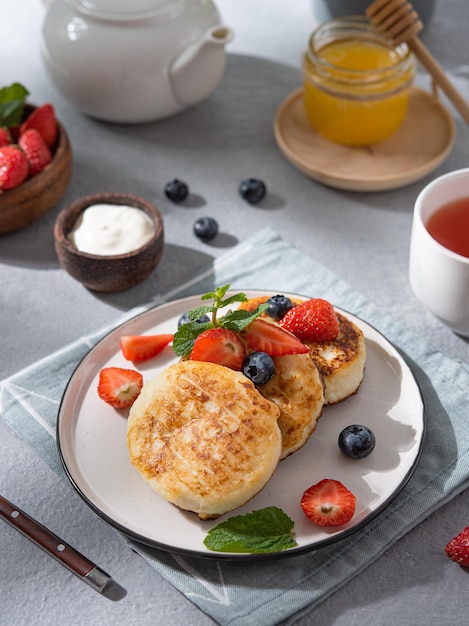 Pannenkoeken met kwark of syrniki met verse bosbessen, aardbeien, honing en zure room op een blauwe achtergrond met een kopje thee en een waterkoker Het concept van gezond en lekker ontbijt