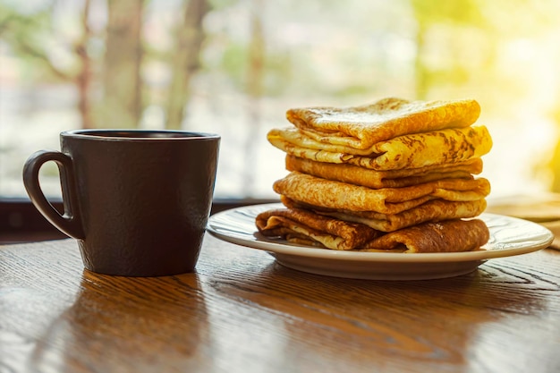 Pannenkoeken op een houten tafel op de achtergrond van een raam Traditioneel Russisch ontbijt Pannenkoeken in de stralen van de felle zon tegen de achtergrond van de natuur van het bos