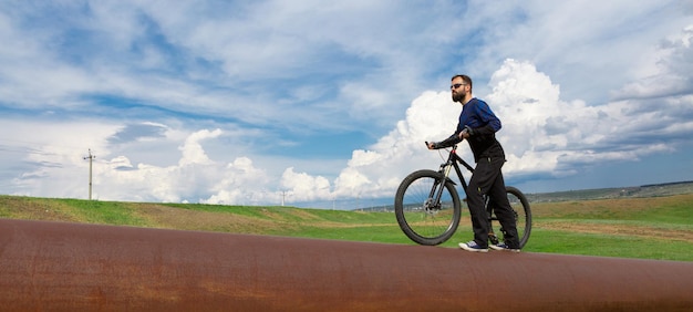 Панорама Бородатый велосипедист на горном велосипеде на ржавой трубе зеленая трава голубое небо.