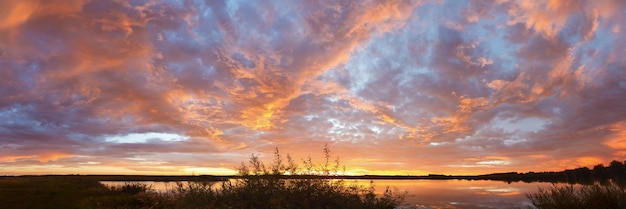 Panorama of a bright colorful sunset over a lake with textured clouds reflected in the water