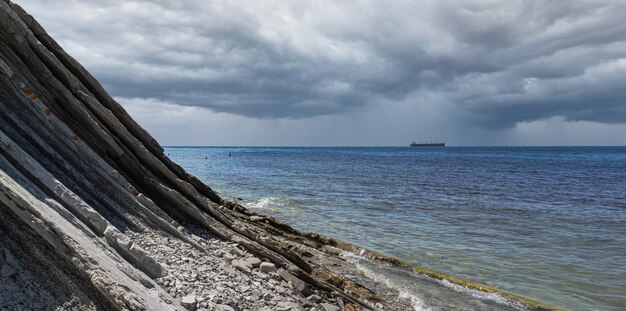 写真 曇りの海のパノラマ。雨天時の岩のふもとにある絵のように美しい石造りの野生のビーチ