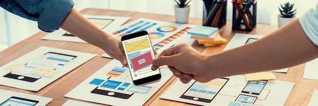 紙のワイヤーフレーム インターフェイス上のモバイル アプリの UI および UX デザインをブレインストーミングしているフロントエンド開発者チームのパノラマ写真 ユーザー フレンドリーな UI デザインを計画しているユーザー インターフェイス開発チーム 精査