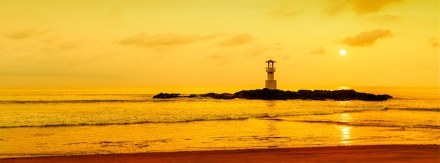 Panorama view of lighthouse on small rock island with orange sky and dramatic clouds at sunset. Taken in south of Thailand.