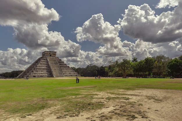 Panorama con la piramide del complesso archeologico di chichen itza in messico circondata dalla vegetazione naturale sotto un cielo di nuvole imbarazzate e alcuni turisti che la ammirano estasiati.