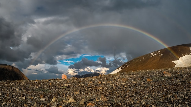 Panorama with a rainbow and a small tourist house in the mountains. Atmospheric alpine landscape with snowy mountains with rainbow in rainy and sunny weather.