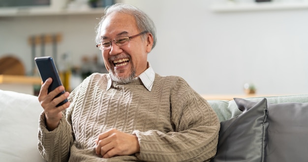 Панорамный счастливый пенсионный пожилой мужчина, сидящий на диване в гостиной, использует мобильный телефон