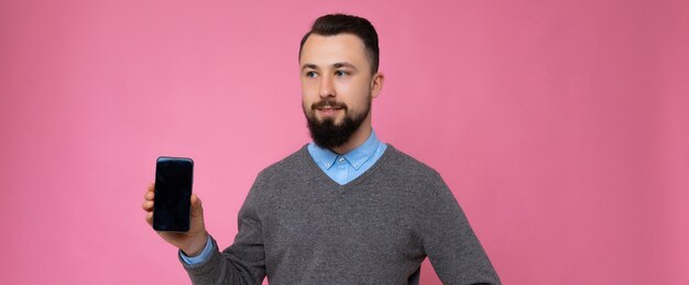 Panoramic photo of handsome happy cool young brunette unshaven man with beardwearing stylish grey sweater and blue shirt standing isolated over pink background wall holding smartphone and showing phon