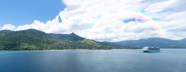 Panoramic photo of Ilha Bela on a sunny day with a ship anchored In the background a rainbow