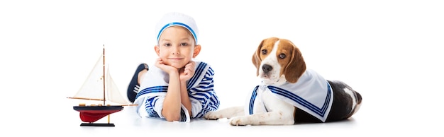 Панорамный снимок мальчика в матросском костюме с игрушечным кораблем и собакой-биглем на белом