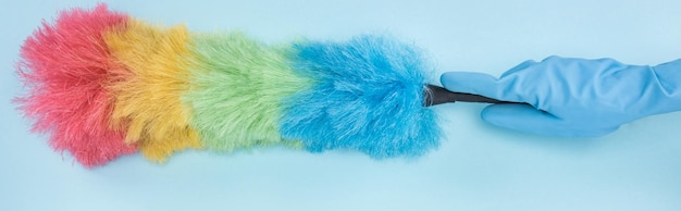 Panoramic shot of cleaner in rubber glove holding rainbow duster on blue background