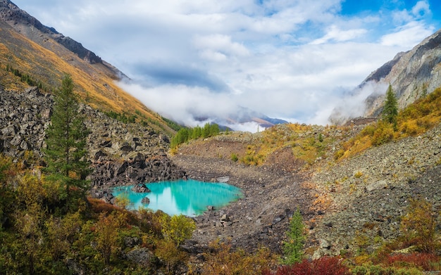 Panoramic view of the autumn mountain, fog over the mountain slopes in the distance, white clouds filling the mountain gorge. A clear blue mountain lake and a bright autumn forest.