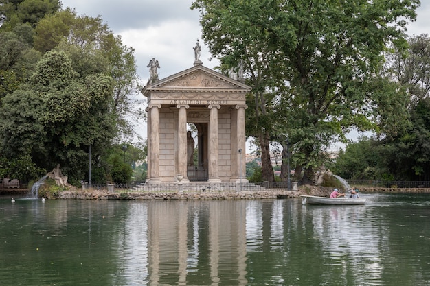 Панорамный вид на храм Асклепия (Tempio di Esculapio) и озеро в общественном парке Вилла Боргезе. Летний день и голубое небо