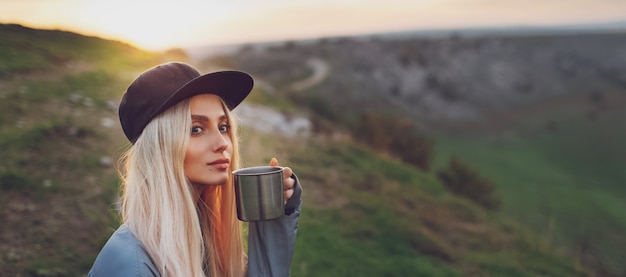 Panoramisch portret van jong blond meisje met stalen beker op de top van de heuvels bij zonsondergang.