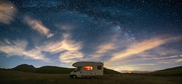 Panoramische nachtelijke hemel over de hooglanden van campo imperatore, abruzzo, italië. de melkwegboog en sterren boven verlichte kampeerauto. kampeervrijheid in een uniek heuvellandschap.