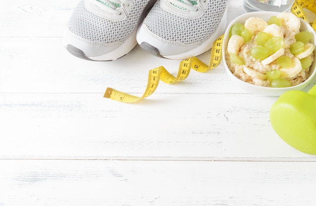 Foto pap met druiven, plakjes banaan, groene halters, sneakers, meetlint op een bovenaanzicht van een wit houten oppervlak