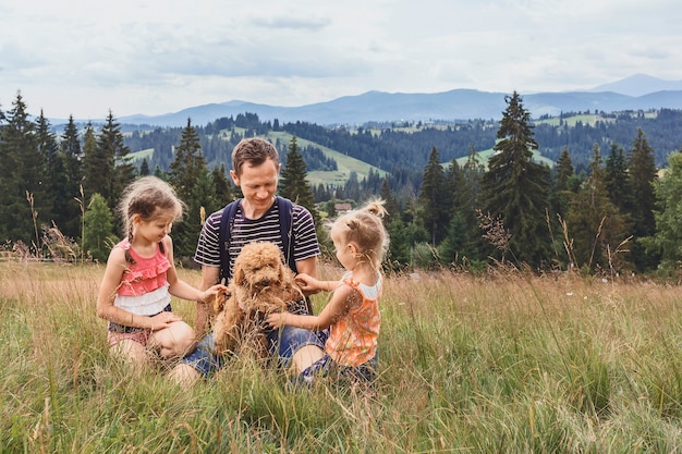 Papa met twee kleine dochters en een hond op een open plek op een achtergrond van bergen