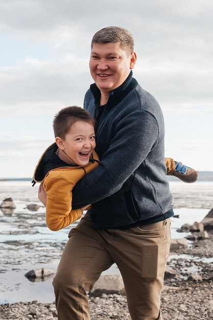 Papa speelt met zijn zoon op de rivieroever. Een mooie vader en zoon hebben plezier met spelen en lachen op een zonnige dag. Het kind ligt in de armen van zijn vader. Buitenrecreatie.