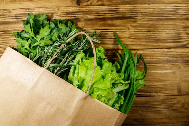 Бумажный пакет с зеленым луком, листьями салата, розмарином и петрушкой на деревянном столе Вид сверху Концепция здорового питания и продуктовых магазинов