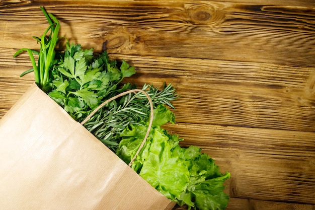 Paper bag with green onion rosemary lettuce leaves and parsley on wooden table Top view Healthy food and grocery shopping concept
