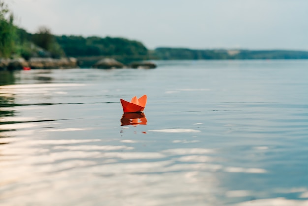 A paper boat sails by the river in the summer. It has an orange color and floats downstream along the shore