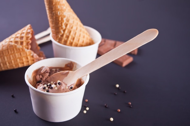 Paper bowls of chocolate ice cream with waffle cones and wooden spoons on the black background