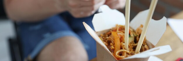 Paper box with chinese noodles standing against background of man with mobile phone in hands closeup