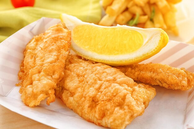 Paper box with tasty fried fish and lemon slice on table closeup