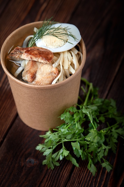 Paper cup noodles with chicken and eggs on wooden background with herbs