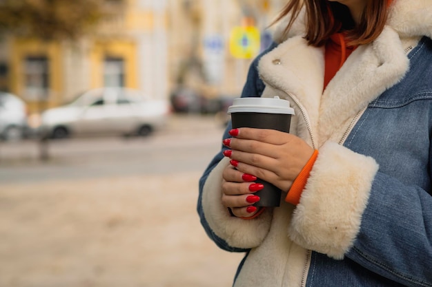 Paper cup with coffee in hands of unrecognizable woman standing in city street Body part of female in warm clothes with bright manicure holding hot drink