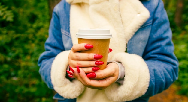 Paper cup with coffee in hands of unrecognizable woman standing in forest Body part of female in warm clothes with bright manicure holding hot drink