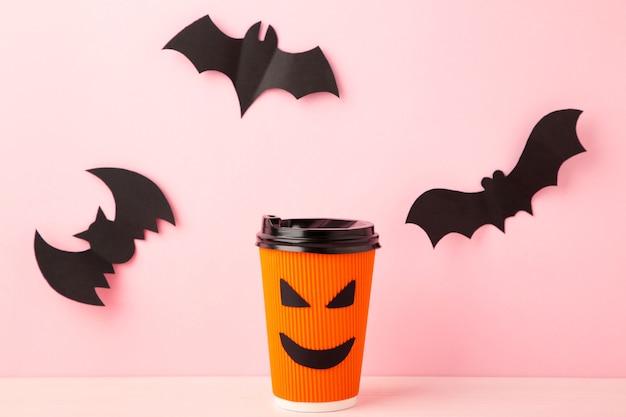 Paper cup with Halloween face in the pink pastel surface with halloween bats