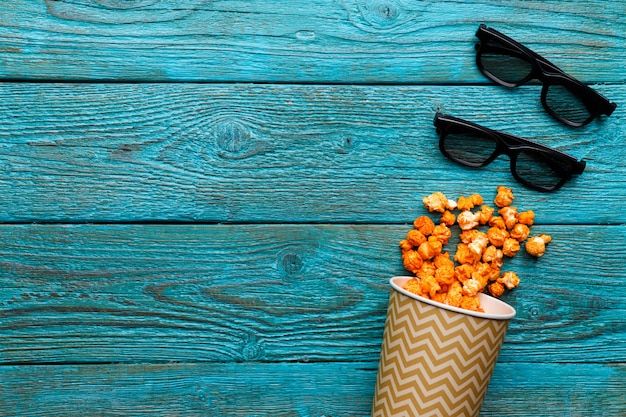 Paper cup with popcorn and 3D glasses on blue background. Top view.