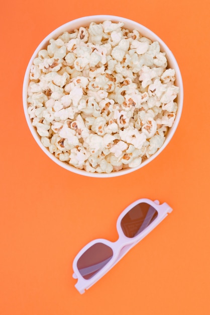 Paper cup with popcorn and 3D glasses on orange background, top view. Flat lay.
