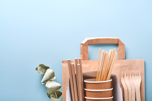 紙コップ、皿、バッグ、木製フォーク、ストロー、ファーストフードコンテナ、水色の背景に木製のカトラリー。エコクラフト紙食器。リサイクルと食品配達の概念。モックアップ。上面図