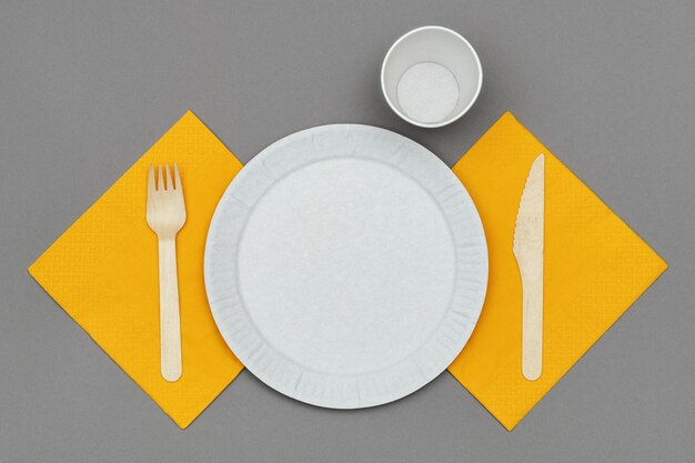 Paper dish and cup, wooden fork and knife on orange napkin on gray background, top view. Set of eco friendly disposable tableware from natural material.