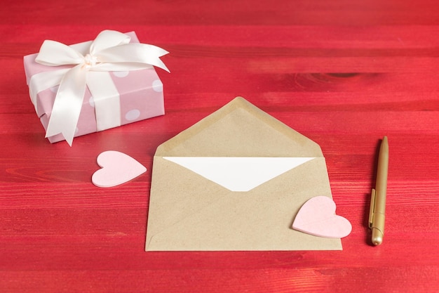 Бумажный конверт с подарочной упаковкой и розовыми сердечками на красном деревянном фоне