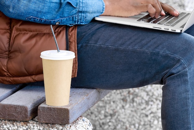 Paper glass with a drink standing on a bench next to a man\
sitting with a laptop