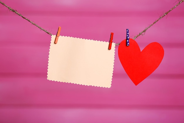 Paper hearts and empty sheet hanging on cord against pink wooden background