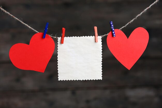 Paper hearts and empty sheet hanging on cord against wooden background